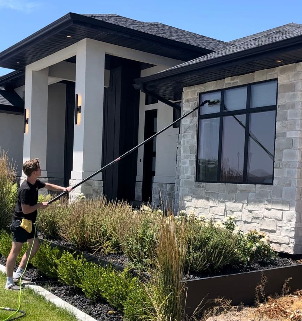 Man using a long pole to wash windows on a modern house with stone exterior and landscaped garden.